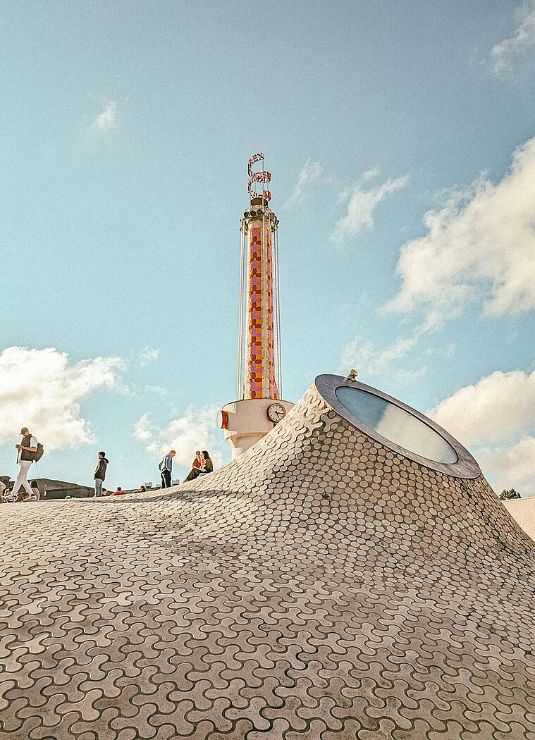 Museo ng Amos Rex sa Helsinki na may tanawing bubong na parang alon at bilugang skylight, may Tore ng Lasipalatsi sa likuran at mga bisita sa liwasan.
